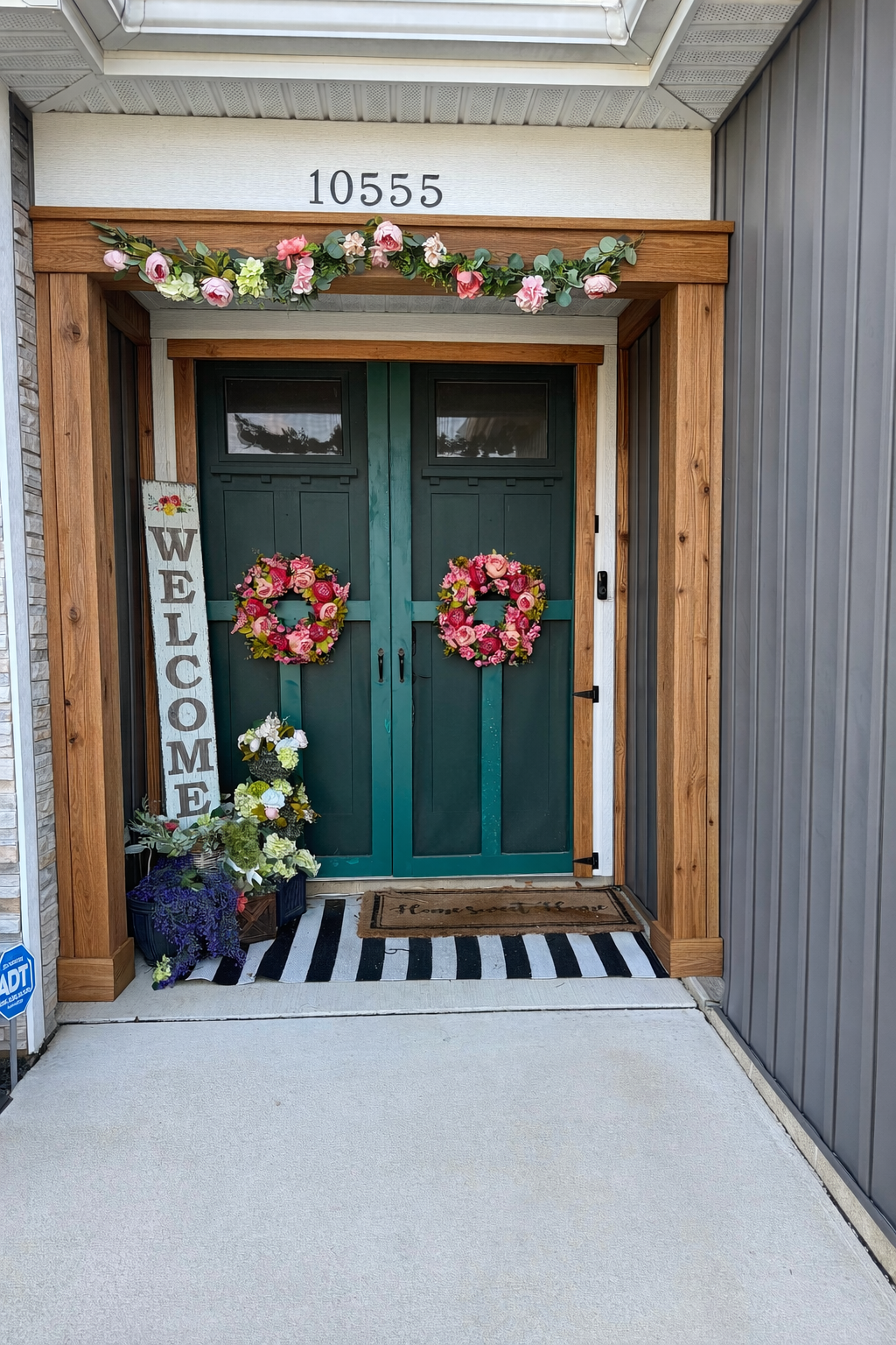Front entryway after — deep teal double doors styled with floral garland, wreaths, and welcome sign